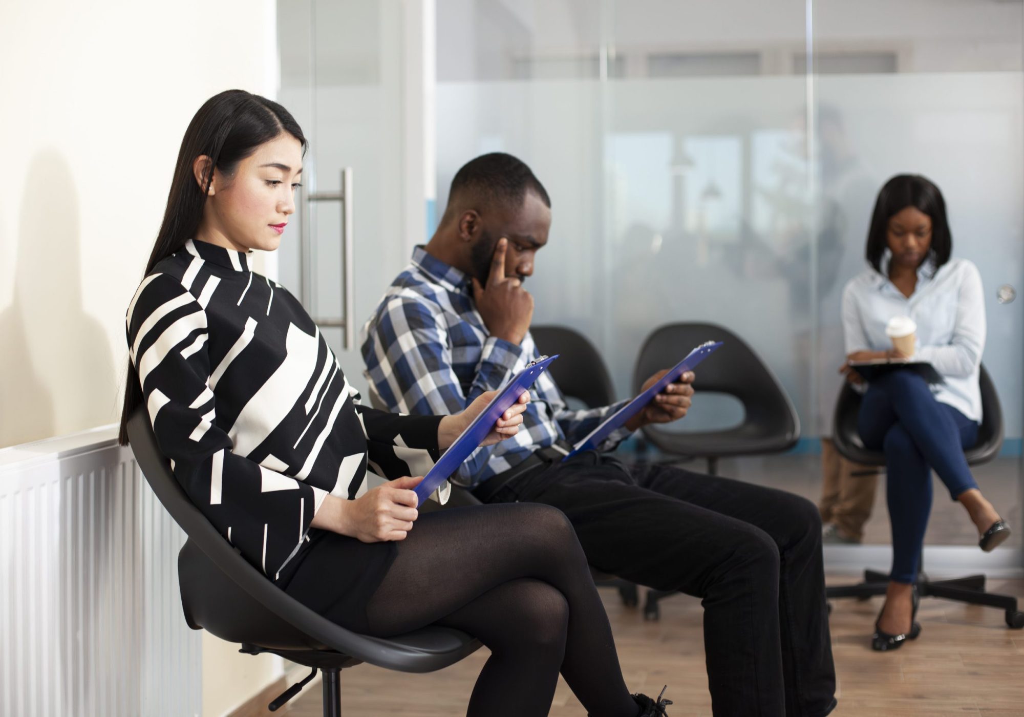 Multiethnic job candidates gather in office lobby for HR meeting, reviewing recruitment documents and holding resumes. Young applicants wait in line for interviews, reading hiring materials.